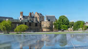 Le Château des Ducs de Bretagne à Nantes, en Loire-Atlantique, se reflète dans un miroir d'eau au premier plan, sous un ciel bleu clair, avec des arbres verts complétant le paysage urbain.