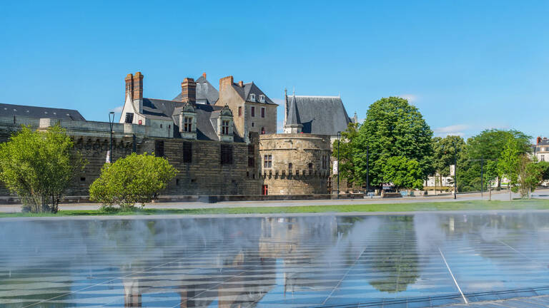 Le Château des Ducs de Bretagne à Nantes, en Loire-Atlantique, se reflète dans un miroir d'eau au premier plan, sous un ciel bleu clair, avec des arbres verts complétant le paysage urbain.