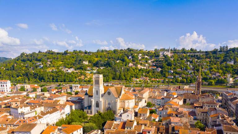 Une vue aérienne d'Agen, en Lot-et-Garonne, avec la Cathédrale Saint-Caprais dominant les toits de la ville, bordée par des collines verdoyantes et le Lot en arrière-plan, sous un ciel bleu parsemé de nuages blancs.