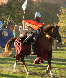 Un chevalier en armure, tenant un drapeau rouge, monté sur un cheval au galop lors d'une reconstitution historique à la Fête Médiévale de Monflanquin en Lot-et-Garonne, avec d'autres drapeaux en arrière-plan sous un soleil couchant.