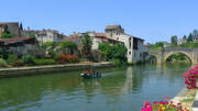 Une balade en bateau sur la Baïse à Nérac, en Lot-et-Garonne, avec les vieilles maisons de pierre et un pont en arc qui bordent la rivière, sous un ciel bleu clair.