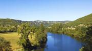 La paisible Vallée du Lot en Lot-et-Garonne, avec la rivière sinuant entre des collines verdoyantes et boisées, sous un ciel bleu clair.