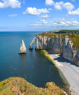 Vue aérienne de la Falaise d'Aval à Étretat, en Normandie (et non dans la Manche), montrant ses célèbres arches naturelles et l'Aiguille creuse se dressant hors de la mer, avec la plage de galets en contrebas, sous un ciel bleu parsemé de nuages.