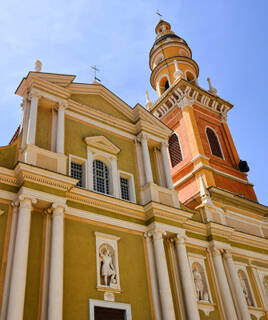 La façade baroque et le clocher de la Basilique Saint-Michel Archange à Menton, avec ses tons ocre et jaunes, ses colonnes et ses statues, sous un ciel bleu clair.