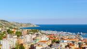 Une vue panoramique sur la ville de Sanremo, en Italie, non loin de la frontière de Menton, avec ses bâtiments le long de la côte, son port de plaisance rempli de bateaux et les collines verdoyantes en arrière-plan, sous un ciel bleu clair.