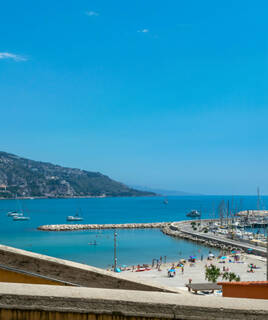 La plage de Menton, avec son port de plaisance rempli de bateaux, une digue et des baigneurs profitant du soleil, le tout sous un ciel bleu clair, avec les montagnes en arrière-plan.