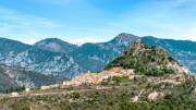 Le village perché de Sainte-Agnès, près de Menton, avec ses maisons aux toits de tuiles rouges et ses façades colorées nichées à flanc de montagne, dominé par les sommets rocheux environnants sous un ciel bleu parsemé de nuages blancs.