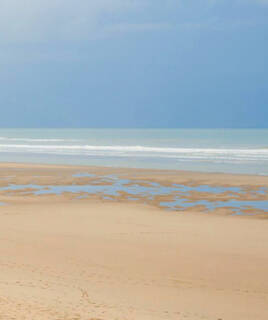 La vaste plage du Touquet-Paris-Plage, près de Merlimont, avec son sable découvert à marée basse et de petites flaques d'eau, menant à l'océan avec des vagues douces sous un ciel nuageux.