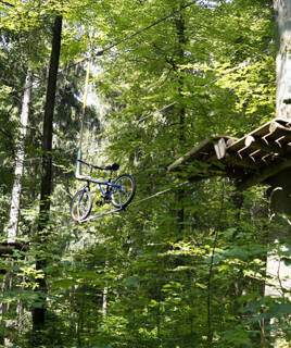 Un vélo suspendu à un câble aérien, faisant partie d'un parcours d'accrobranche ou d'aventure à Adrénaline Parc à Moliets, près de Messanges, entouré d'arbres verts sous la lumière du soleil.