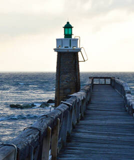 Le phare vert et blanc de la jetée de Capbreton, près de Messanges, s'élève au-dessus des eaux agitées de l'océan, au bout d'un long ponton en bois, sous un ciel nuageux.