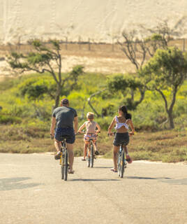Une famille de trois personnes à vélo sur une piste cyclable de la Vélodyssée près de Messanges, avec des dunes de sable et de la végétation en arrière-plan sous un ciel ensoleillé.