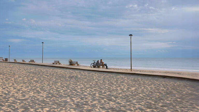 Une longue plage de sable à Montalivet, avec une promenade en bois et des bancs, sous un ciel légèrement nuageux, et une personne assise près d'un vélo au bord de l'océan.