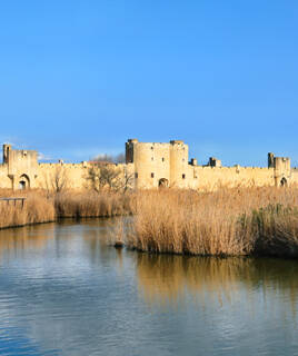 Les remparts imposants de la cité médiévale d'Aigues-Mortes, près de Montpellier, se reflétant dans les eaux calmes des marais environnants, sous un ciel bleu.