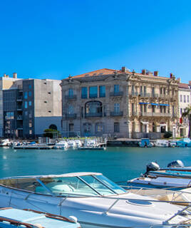 Le port de Sète, près de Montpellier, avec de nombreux bateaux amarrés sur des eaux bleues, bordé par des bâtiments historiques et modernes sous un ciel clair.