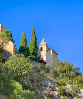 La Chapelle Notre-Dame de Beauvoir à Moustiers-Sainte-Marie, perchée à flanc de montagne, avec son clocher et ses murs de pierre claire, entourée de cyprès et de végétation, sous un ciel bleu éclatant.