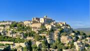 Le village perché de Gordes, dans le Luberon (Provence-Alpes-Côte d'Azur), avec ses maisons en pierre et son église dominant la colline, entouré d'une végétation luxuriante sous un ciel bleu intense.