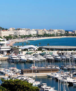 Le port de Cannes, en Provence-Alpes-Côte d'Azur, rempli de yachts et de bateaux de plaisance, avec la célèbre Croisette, sa plage et ses bâtiments emblématiques en arrière-plan, sous un ciel bleu dégagé.