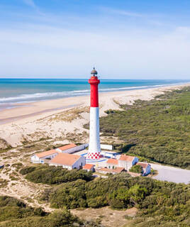 Le Phare de la Coubre, près de La Palmyre, se dresse fièrement sur la côte, avec ses bandes rouges et blanches, surplombant la plage de sable et l'océan Atlantique, entouré de végétation, sous un ciel bleu éclatant.