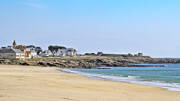 La plage de Penthièvre, dans la Baie de Quiberon, près de Pénestin, avec son sable fin, ses rochers et ses maisons côtières en arrière-plan, sous un ciel bleu clair.