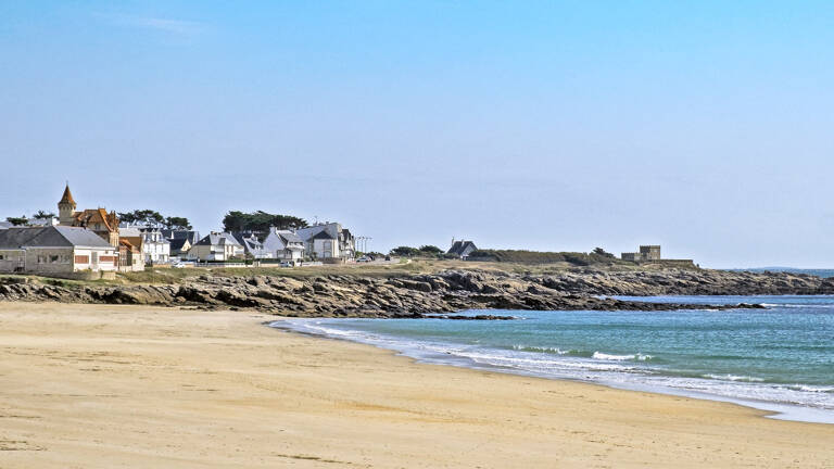 La plage de Penthièvre, dans la Baie de Quiberon, près de Pénestin, avec son sable fin, ses rochers et ses maisons côtières en arrière-plan, sous un ciel bleu clair.