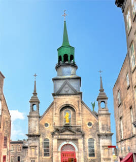 La façade de la Chapelle Notre-Dame de Bon Secours à Pénestin, une église en pierre avec une flèche verte et une statue dorée au-dessus de l'entrée rouge, sous un ciel bleu clair.