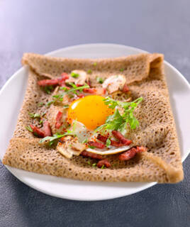 Une galette de sarrasin bretonne, spécialité de Pénestin, garnie d'un œuf au plat, de champignons et de lardons, servie sur une assiette blanche.