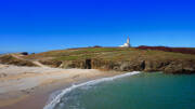 Le phare de Port Navalo dans le Golfe du Morbihan, près de Pénestin, se dresse sur une pointe rocheuse, dominant une plage de sable et les eaux turquoise de l'océan, sous un ciel bleu intense.