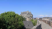 Les remparts de Guérande, près de Pénestin, avec une tour en pierre et des toits d'ardoise visibles derrière les arbres, offrant une vue sur la ville historique sous un ciel bleu clair.