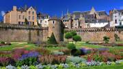 Les jardins à la française des remparts de Vannes, près de Pénestin, avec des parterres de fleurs colorés et des arbustes taillés, dominés par les murs de pierre de la ville médiévale et ses bâtiments historiques sous un ciel bleu éclatant.