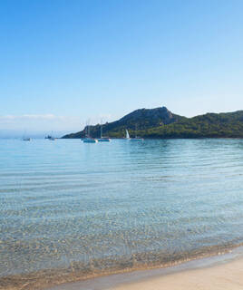 La plage de Notre-Dame à Porquerolles, avec ses eaux cristallines et peu profondes, quelques voiliers au large, et une colline boisée en arrière-plan sous un ciel bleu clair.