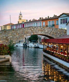 Un canal de Port Grimaud, avec des bateaux amarrés et des bâtiments colorés bordant les rives, dont un restaurant flottant sous un pont en pierre, le tout sous une lumière de crépuscule.