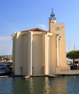 L'église Saint-François d'Assise de Port Grimaud, avec sa façade crème et son clocher, se reflétant dans les eaux du canal, entourée de bateaux amarrés sous un ciel bleu clair.