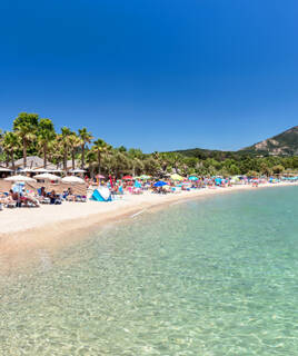 La plage de Port Grimaud, une plage de sable animée avec de nombreux parasols et chaises longues, bordée de palmiers et d'eaux claires, sous un ciel bleu éclatant, avec des montagnes en arrière-plan.