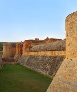La Forteresse de Salses-le-Château, près de Port Leucate, avec ses imposants murs de pierre ocre et ses bastions, entourée de fossés verdoyants, sous un ciel bleu clair.
