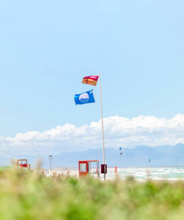 Un mât avec un drapeau rouge "Off Duty" et un drapeau bleu "Pavillon Bleu" flottant sur une plage de Port Leucate, avec des équipements de surveillance en arrière-plan et l'océan avec des kitesurfeurs sous un ciel bleu.