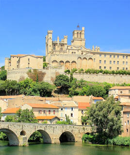 La Cathédrale Saint-Nazaire de Béziers, près de Portiragnes, domine la ville du haut de sa colline, avec un pont en pierre enjambant une rivière au premier plan et des maisons en contrebas, sous un ciel bleu éclatant.