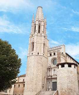 L'église Saint-Félix de Gérone, près de Portiragnes, avec son imposant clocher gothique en pierre et sa façade richement décorée, sous un ciel bleu parsemé de nuages blancs.