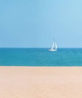 La plage de sable fin de Valras-Plage, près de Portiragnes, avec un voilier blanc naviguant sur les eaux turquoise de la Méditerranée sous un ciel bleu clair.