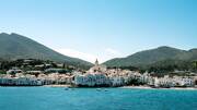 Le village côtier de Cadaqués, en Catalogne, avec ses maisons blanches s'étageant à flanc de colline jusqu'à l'église centrale, bordant une baie aux eaux bleues, le tout encadré par des montagnes arides sous un ciel clair.