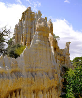 Les Orgues d'Ille-sur-Têt, dans les Pyrénées-Orientales, des cheminées de fées sculptées par l'érosion, se dressant majestueusement avec leur couleur ocre et blanche, entourées de végétation sous un ciel bleu parsemé de nuages blancs.