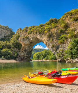 Le Pont d'Arc en Ardèche, une arche naturelle impressionnante surplombant la rivière, avec des kayaks colorés sur la berge et une végétation luxuriante sous un ciel bleu éclatant, invitant aux activités nautiques.