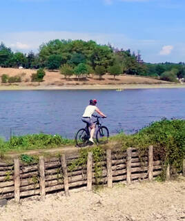 Une personne à vélo sur un sentier longeant une rivière, avec un barrage de bois au premier plan, des arbres en arrière-plan et un ciel partiellement nuageux, évoquant une balade à vélo en bord de rivière.