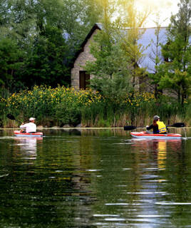 Deux personnes faisant du canoë sur un lac calme, l'une portant un gilet de sauvetage jaune, avec une maison en pierre et de la végétation en arrière-plan, sous un ciel lumineux, illustrant une activité nautique près de Saint-Gilles-Croix-de-Vie.