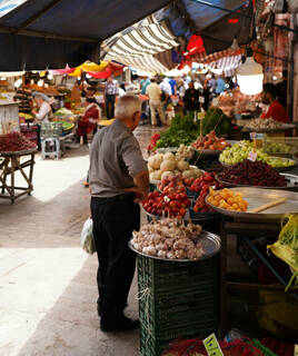 Un marché animé à Saint-Gilles-Croix-de-Vie, avec un homme au premier plan devant un étal de fruits et légumes frais, dont des têtes d'ail, sous des auvents colorés.