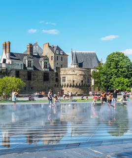 Le miroir d'eau de Nantes, avec ses reflets, devant le Château des Ducs de Bretagne, sous un ciel bleu, avec des personnes profitant du lieu.