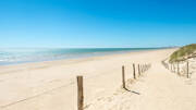 Des planches de bois et une clôture délimitent un chemin de sable menant à une vaste plage dorée à Saint-Hilaire-de-Riez, avec l'océan atlantique calme sous un ciel bleu éclatant.
