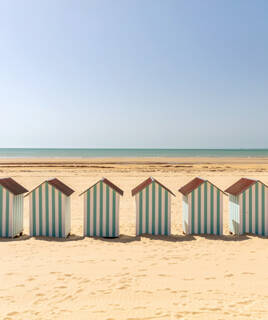 Sept cabines de plage rayées vertes et blanches alignées sur le sable de la Plage des Demoiselles à Saint-Hilaire-de-Riez, avec l'océan calme en arrière-plan sous un ciel dégagé.