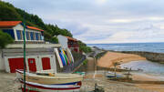 Le charmant petit port de Guéthary, près de Saint-Jean-de-Luz, avec des bateaux de pêche sur la plage et des maisons traditionnelles basques à flanc de colline, et la mer agitée en arrière-plan sous un ciel nuageux.