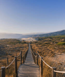 Un long sentier en bois avec des cordes pour balustrade, traversant les dunes de sable et la végétation sèche du Sentier de la Dune à Saint-Jean-de-Monts, s'étendant vers l'horizon sous un ciel clair.