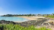 Plage de Port Mer à Cancale, près de Saint-Malo, avec son sable doré et son eau turquoise parsemée de bateaux, bordée de rochers et d'une végétation luxuriante, avec le village en arrière-plan sous un ciel bleu.